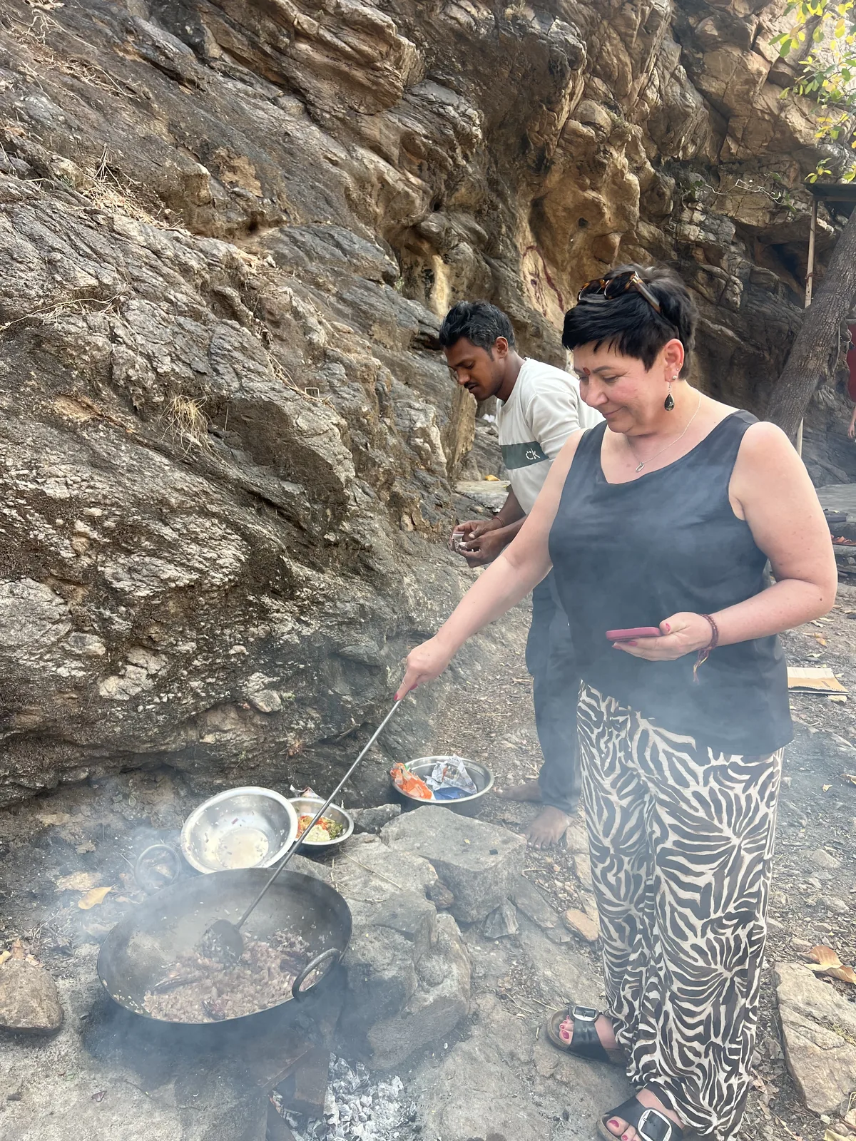 A visitor learns a cooking technique at a fire near the cave